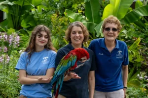 Heather, the volunteer services manager stands with her mother and daughter. Heather holds out her hand with a red, green, and blue macaw perched on it.