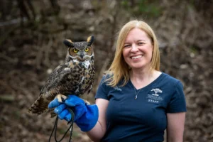Brea, a Senior Education Specialist stands with an Owl perched on her gloved hand. She had blonde hair and wears a navy blue staff uniform shirt.