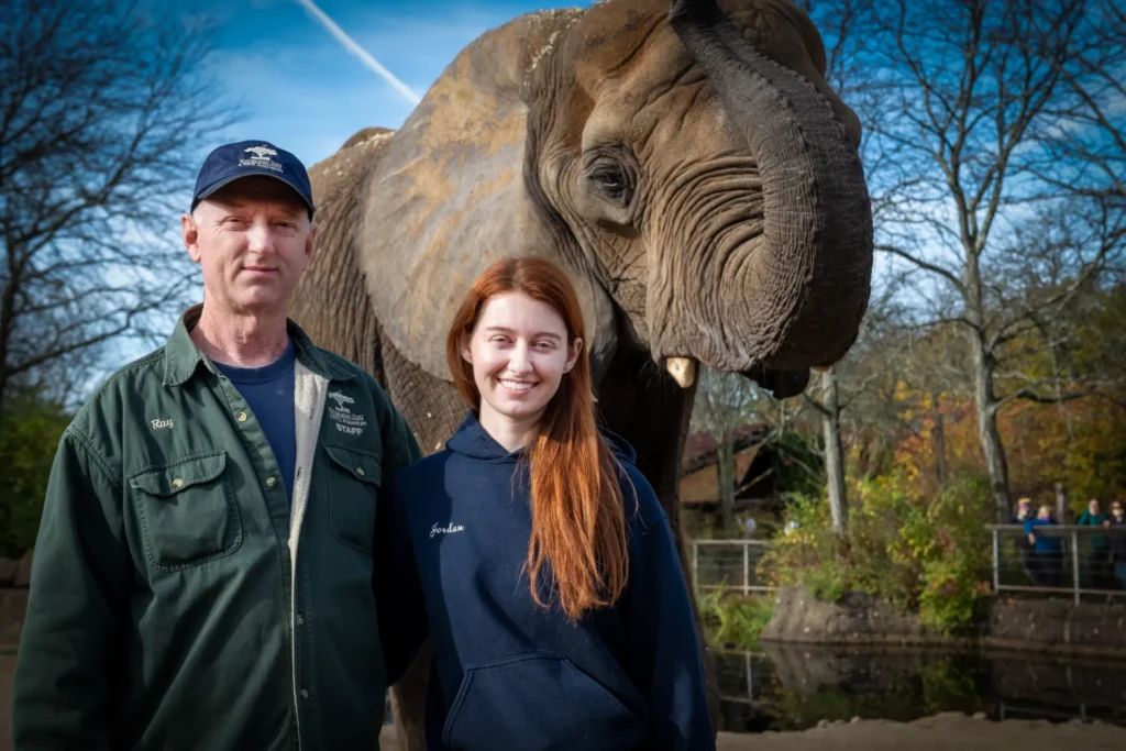 ray and jordan in front of an elephant. ray is wearing a zoo hat and jacket and jordan is wearing a blue zoo hoodie. the elephant has a raised trunk.