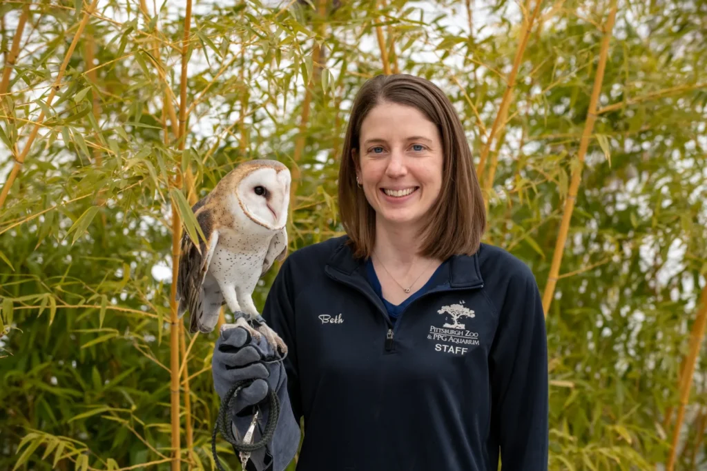 beth with an ambassador animal perched on her gloved hand. beth has straight brown hair and is wearing a dark zoo staff zip up. she is standing outside in front of bamboo.