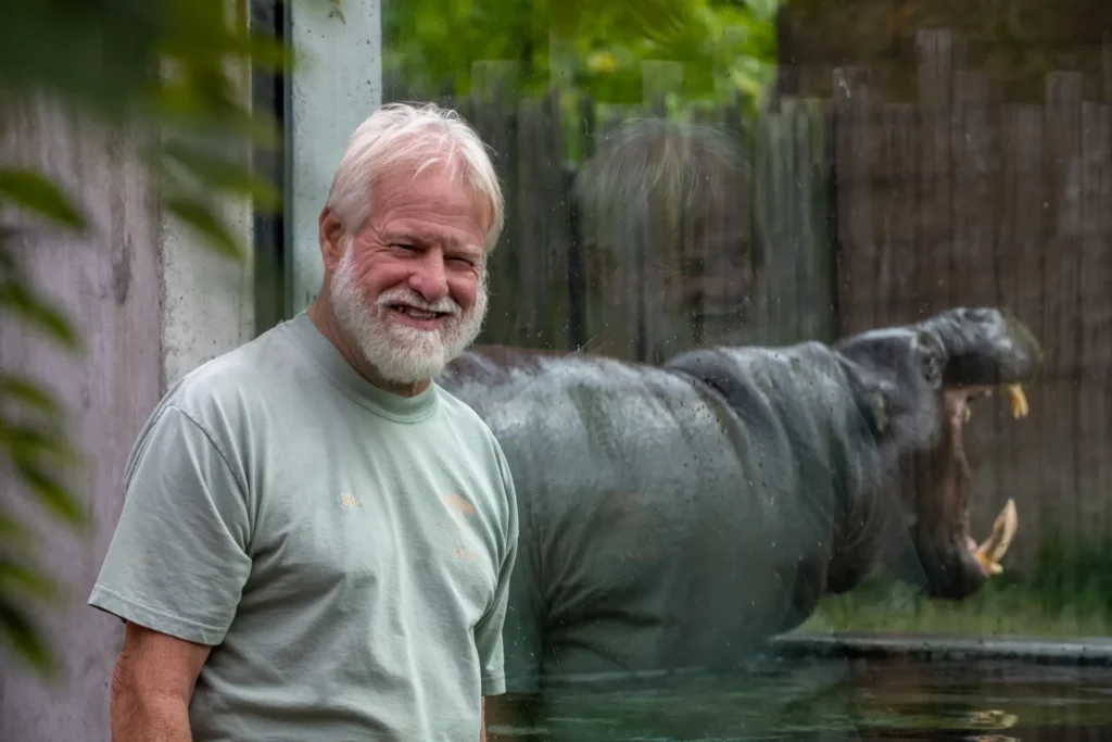 mo in front of the pygmy hippo habitat. mo has white facial hair and a gray shirt. the hippo has his mouth open in the background.