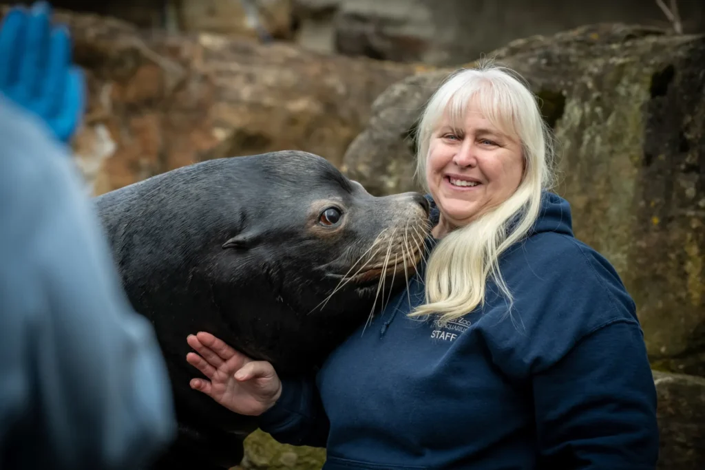 judy with a sea lion. judy has light blonde hair and the sea lion is close to her face. she has a hand on the se lion's neck.
