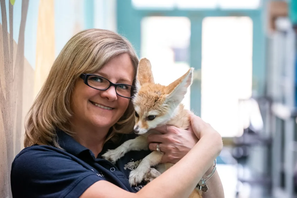 lori holding a fennec fox. lori has short blonde hair and is wearing glasses and a blue shirt. she is holding the fennec fox close to her face.
