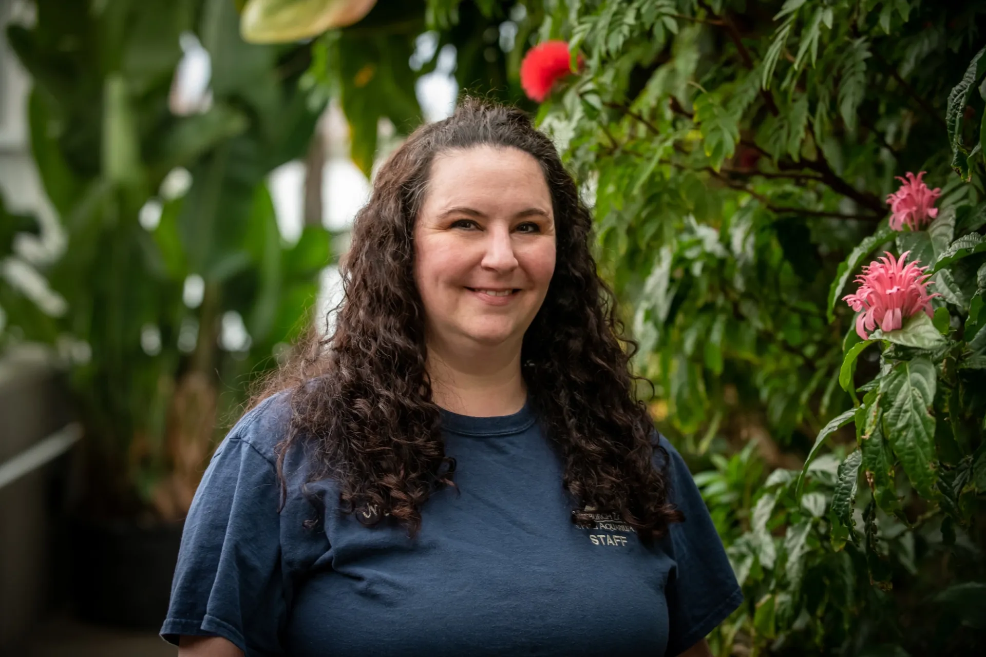 nicole in the rainforest section of the aquarium. nicole has curly brown hair and is wearing a blue zoo staff shirt. she is standing in front of tropical flowers.